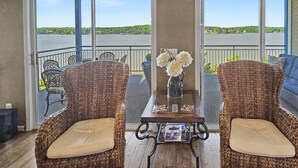 Een balkon met rieten stoelen, een tafel met een bloevas, en uitzicht op een meer.