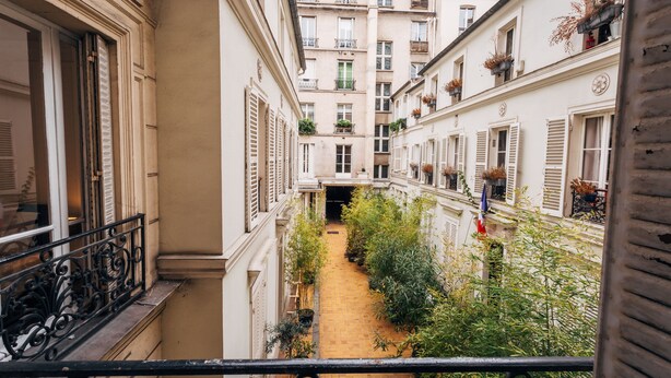 Une ruelle étroite entre deux immeubles, avec des balcons, des plantes en pot et un drapeau.