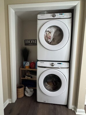 Een wasserij met twee gestapelde Samsung wasmachines, een plank met schoonmaakmiddelen, en een bordje met de tekst 'HOME Gewoon sinds 1928'.