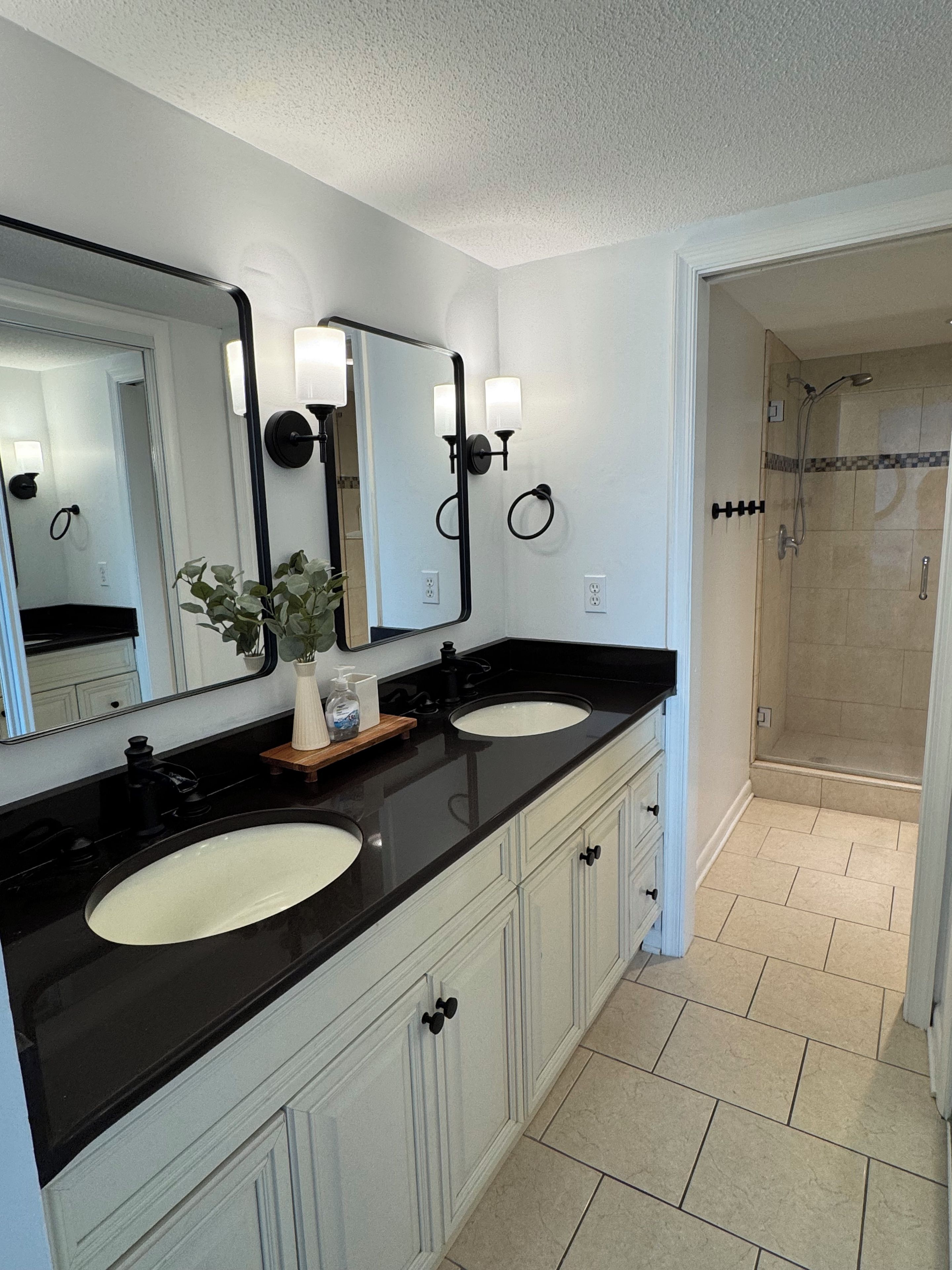 A bathroom with two sinks, a large mirror, wall sconces, a potted plant, and a shower area.