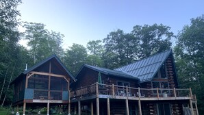 Une cabane en rondins dotée d’un balcon et d’une terrasse couverte, entourée d’arbres.