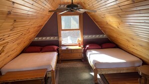 Um quarto de sótão acolhedor com duas camas de solteiro, um ventilador de teto de madeira e uma janela com vista para uma cerca de madeira.