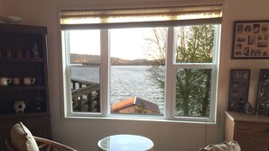 Ein Zimmer mit großem Fenster und Blick auf ein Gewässer, zwei Korbstühle, ein kleiner runder Tisch und eine Holzkommode mit einer Sammlung gerahmter Fotos.