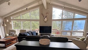 Ein geräumiges Wohnzimmer mit einer großen, schwarzen Unterhaltungsanlage, einem aufgehängten Hirschkopf und großen Fenstern mit Blick auf Bäume und eine Terrasse.