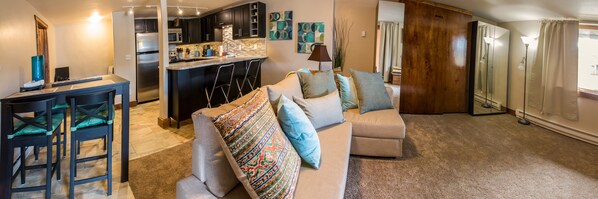 A modern living room with a beige sofa, a dining area with a table and chairs, a kitchen with dark cabinets and a stainless steel refrigerator, and a large window with curtains. - 500 Ft. From Slope! Remodeled Studio (Steamboat Springs)