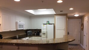 A kitchen with white cabinets, a granite countertop, a refrigerator, a microwave, a coffee maker, and a toaster. There are four stools at the bar area.
