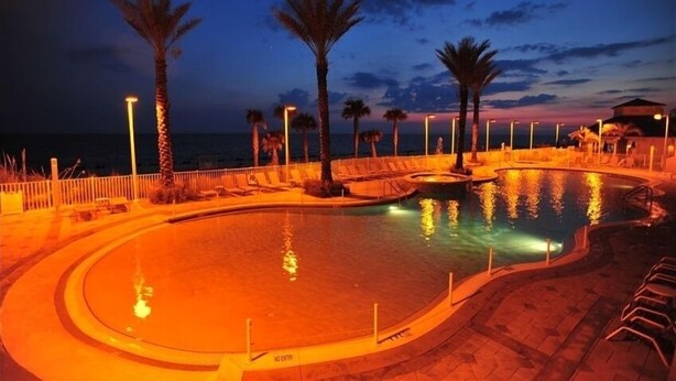 A pool area with a curved design, palm trees, and a view of the sea at dusk.