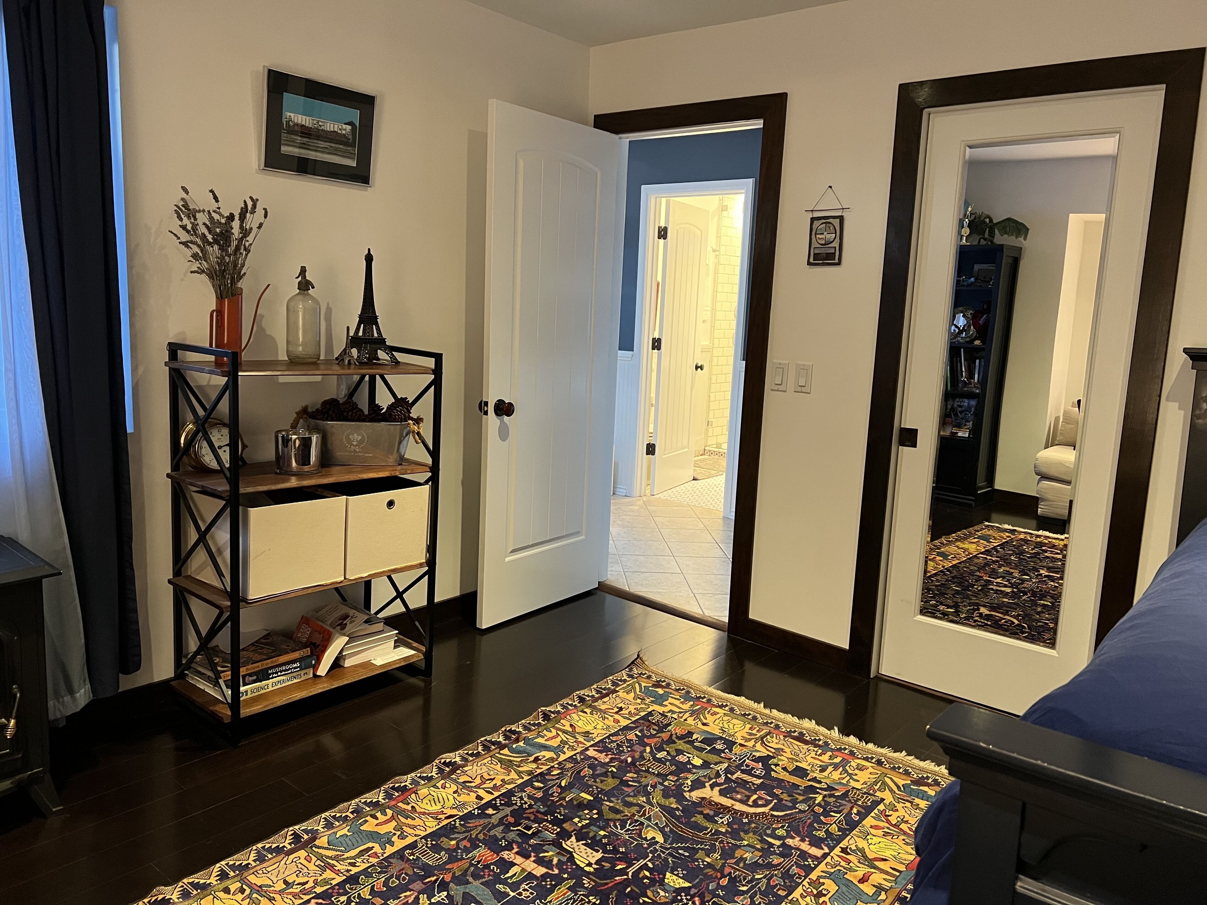 A room with a dark wooden floor, a patterned rug, a bookshelf with decorative items, and a door leading to another room.
