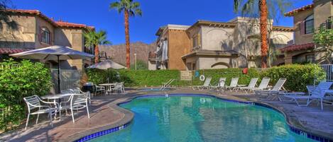 A swimming pool with lounge chairs and umbrellas, surrounded by palm trees and a mountainous backdrop.