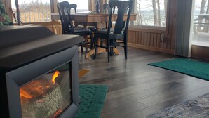 Una habitación acogedora con una estufa de leña, una mesa de comedor con sillas, ventanas amplias y vistas a árboles.
