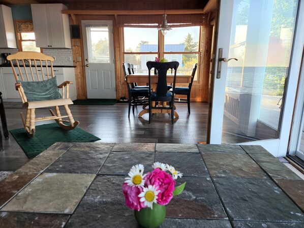 A wooden interior with a dining table and chairs, a rocking chair, and a green rug.