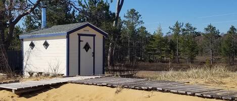 Eine kleine Holzhütte mit Tür und Fenstern, umgeben von Sand und Bäumen.