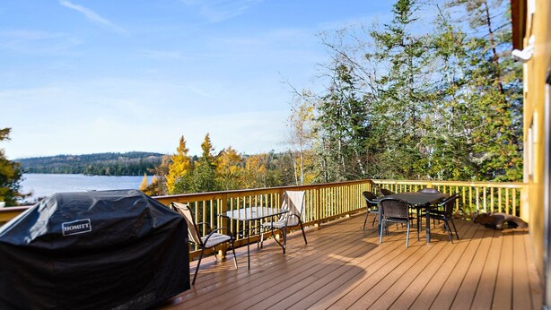 Una terraza de madera con mobiliario exterior, una parrilla y vistas a un lago y árboles.