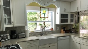 Une cuisine avec des meubles blancs, une cuisinière à gaz, un évier et une fenêtre décorée de fleurs.
