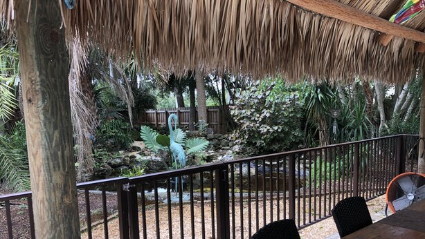 Un espace extérieur abrité, doté d’une table et de chaises en bois, d’une rambarde en métal et offrant une vue sur un jardin tropical agrémenté d’un élément aquatique et d’une statue.