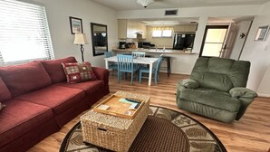 Un salon avec un canapé rouge, un fauteuil vert, une table basse et un coin repas avec des chaises bleues.
