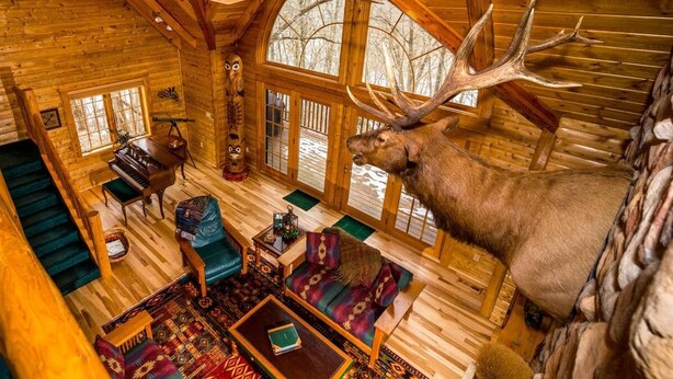 A log cabin interior with a grand staircase, a piano, a large deer head mounted on the wall, and a cozy seating area. - Wildlife Surrounds! Enjoy Fireplace, Pool Table, Fire Pit, Piano. Near Marina! (Galena)