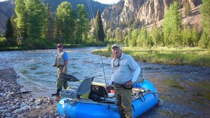 Deux personnes se tiennent à côté d’un radeau gonflable bleu sur la rive d’une rivière, avec une chaîne de montagnes en arrière-plan.