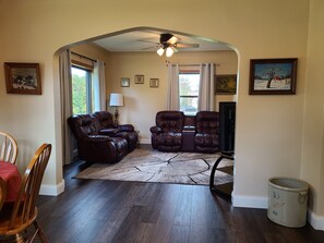 Een woonkamer met donker houten vloeren, leren relaxfauteuils, een eettafel met stoelen, een plafondventilator en lijstwerken aan de muren.