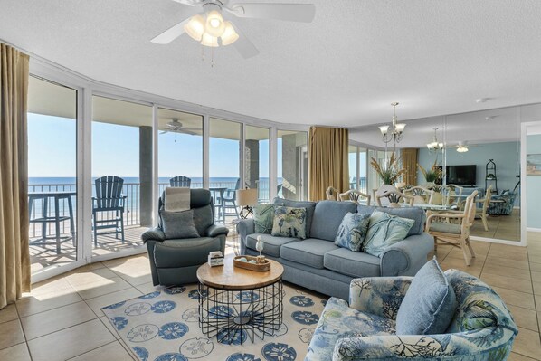 A spacious living area with a large window overlooking the ocean, a ceiling fan, and a dining area with wicker furniture.