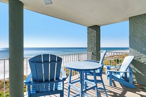A covered outdoor seating area with blue Adirondack chairs and a small table, overlooking a beach with a clear view of the ocean.