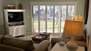 Una sala de estar con televisor, mueble blanco para entretenimiento y un ventanal con persianas.