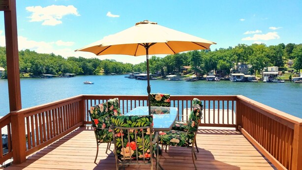 Una terraza de madera con una mesa, sillas, un paraguas y vistas a un lago con barcos y casas al fondo.