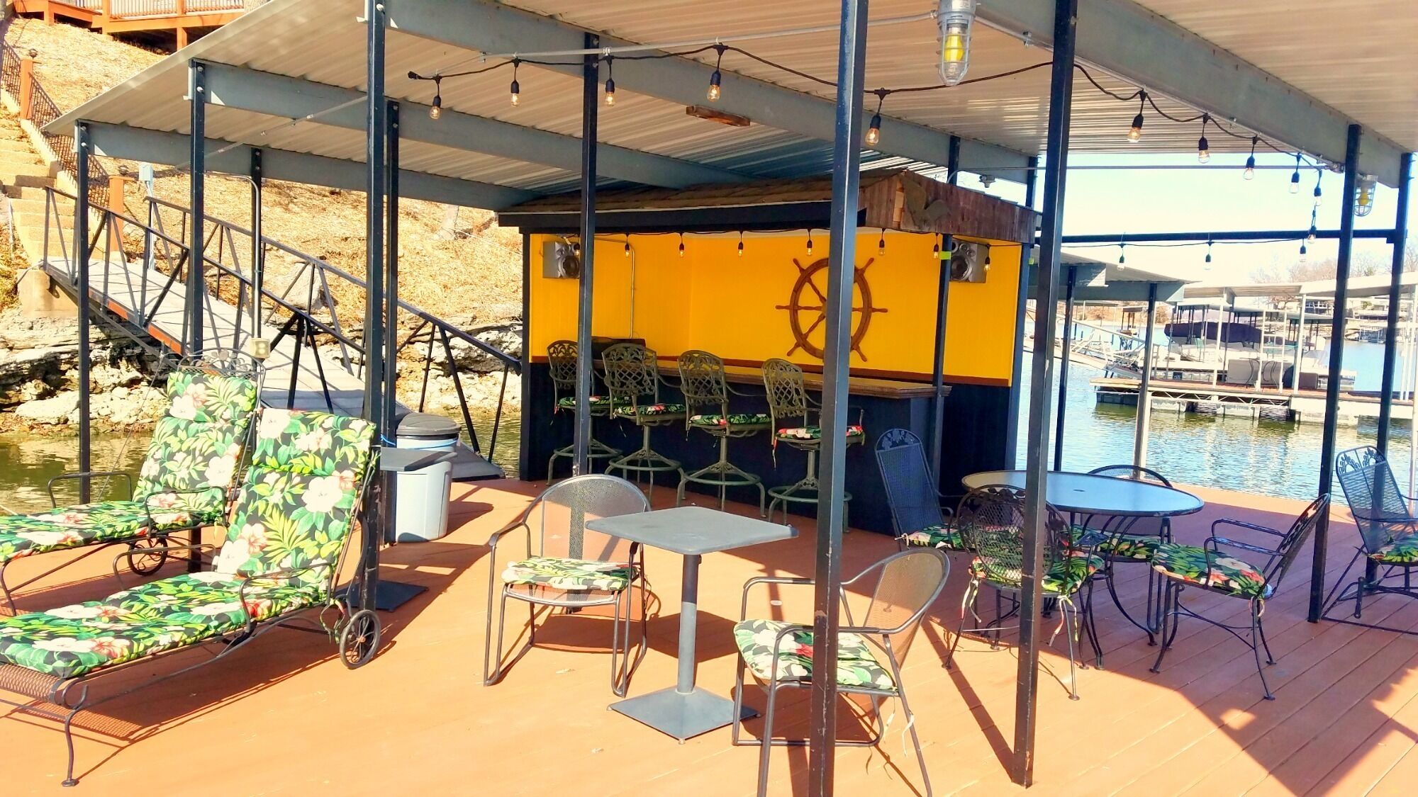 A covered outdoor seating area with chairs and tables, a staircase leading to a higher level, and a view of the water and boats in the background.