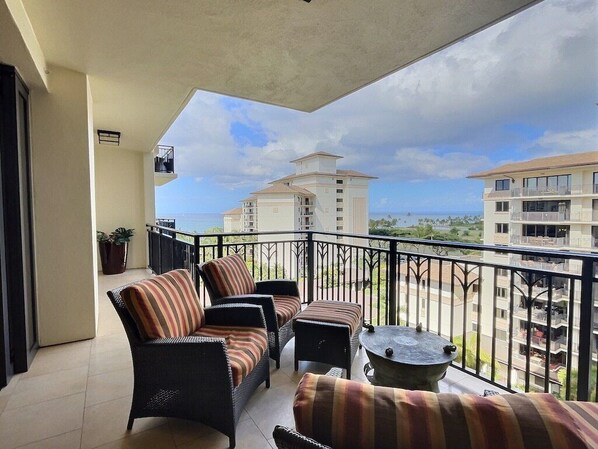 A balcony with wicker furniture, a round table, and a view of neighboring buildings and the ocean.