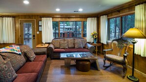 A cozy living room with a sofa, armchair, and coffee table, wooden walls, and large windows with curtains. - Buss Stop (Wawona)