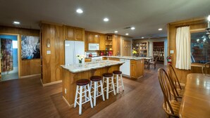 A spacious kitchen with wooden cabinets, a large island with bar stools, a dining area with a wooden table and chairs, and a view of a forest through a window. - Buss Stop (Wawona)
