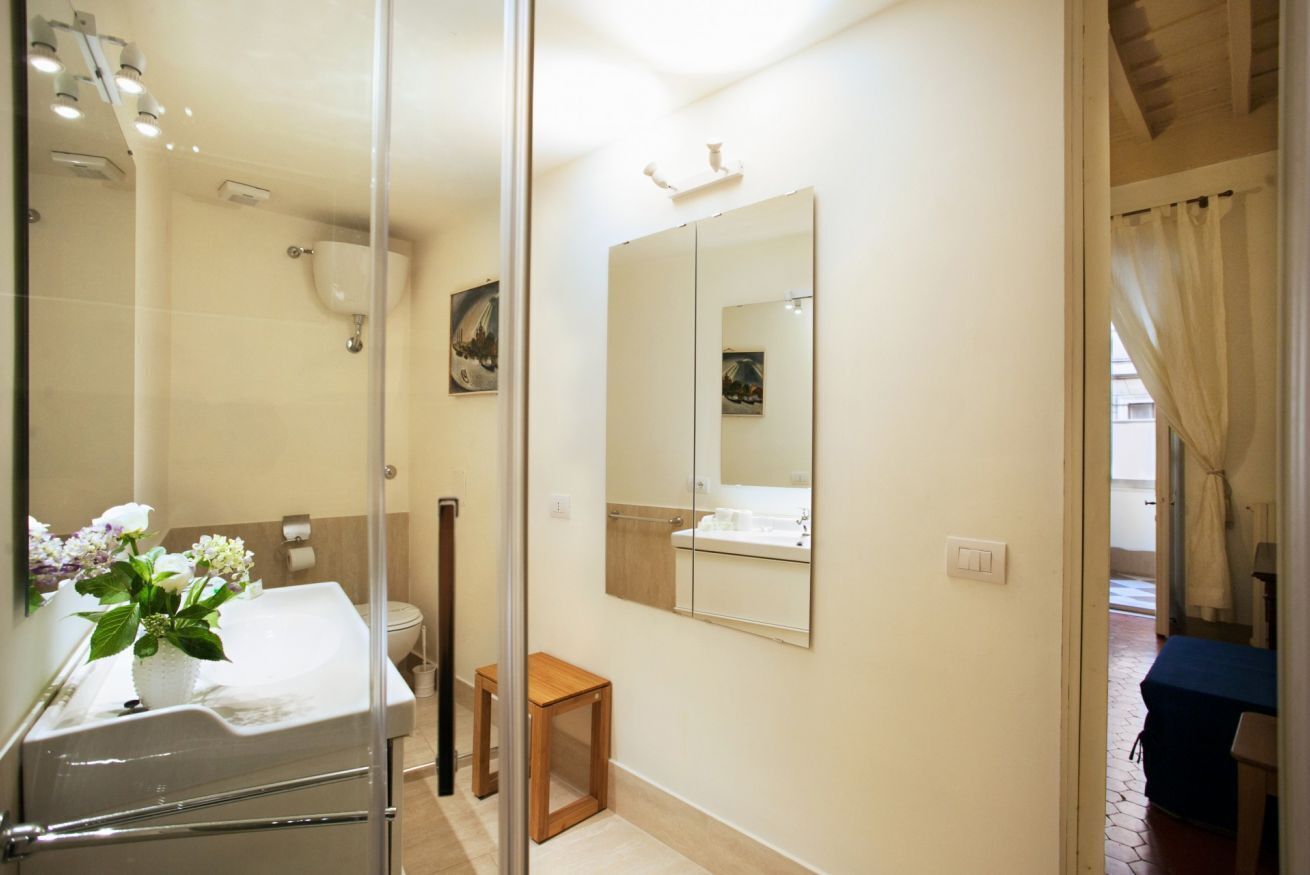 A bathroom with a glass shower enclosure, a white sink with a vase of flowers, a wooden stool, a large mirror, and a toilet.