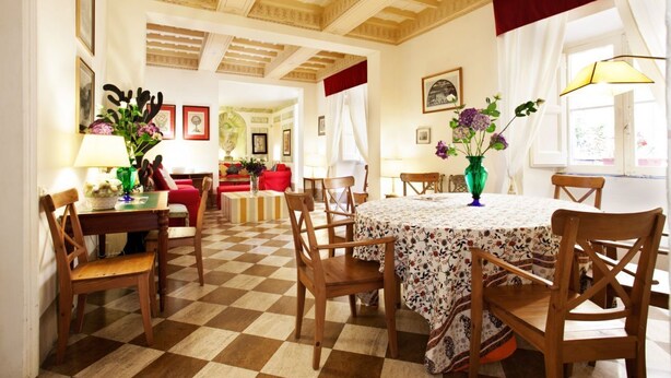 Zona pranzo con pavimento a scacchi, sedie di legno, un tavolo rotondo con tovaglia floreale e un lampadario.