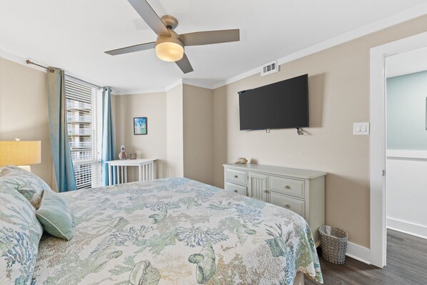 A bedroom with a bed, a ceiling fan, a TV mounted on the wall, a small table, and a window with blinds.