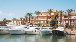 A marina with several yachts docked, surrounded by multi-story buildings with balconies and palm trees.