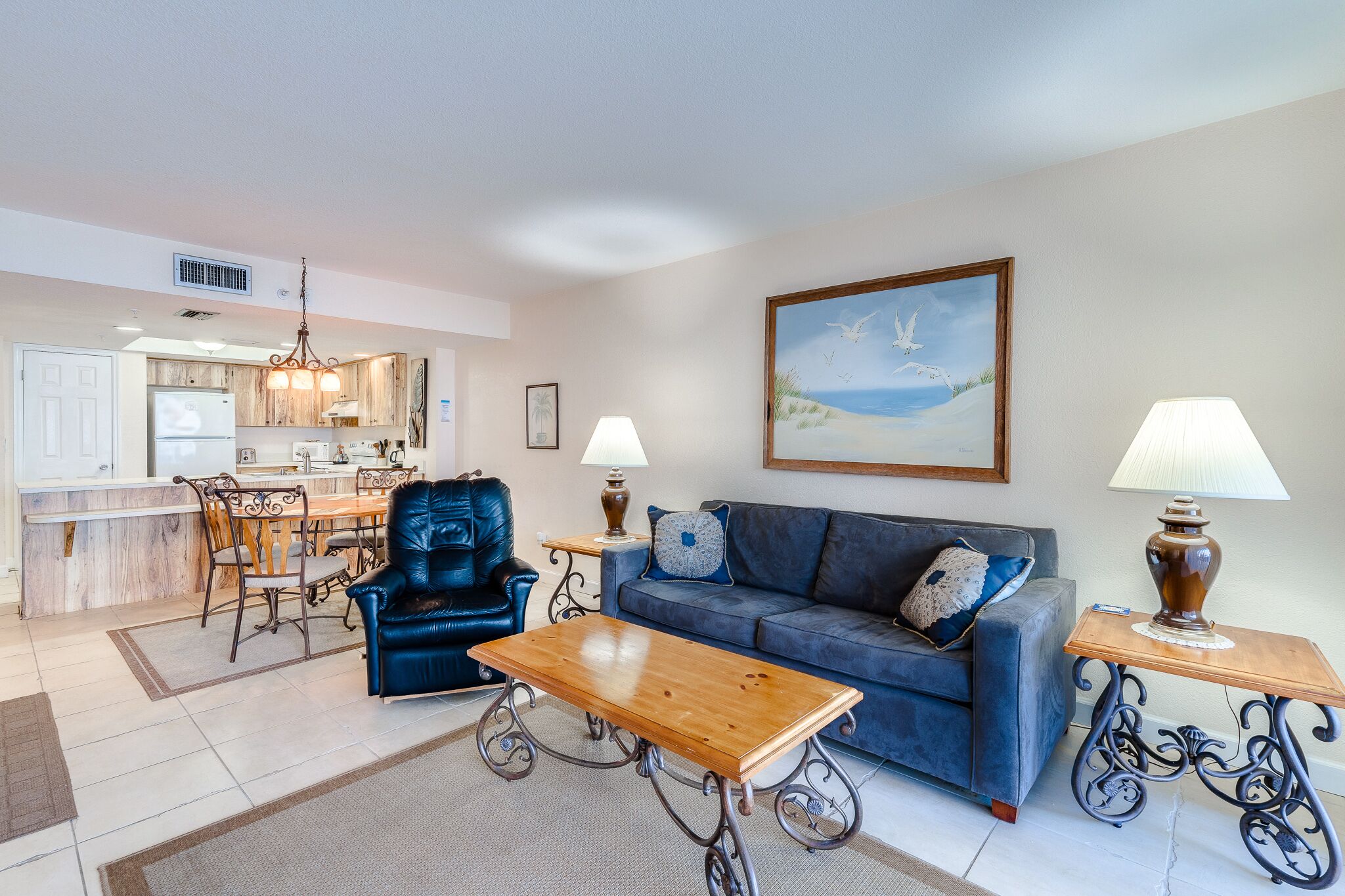 Un salon avec un canapé bleu, une table basse en bois, un fauteuil en cuir noir et deux lampes sur des tables d’appoint.