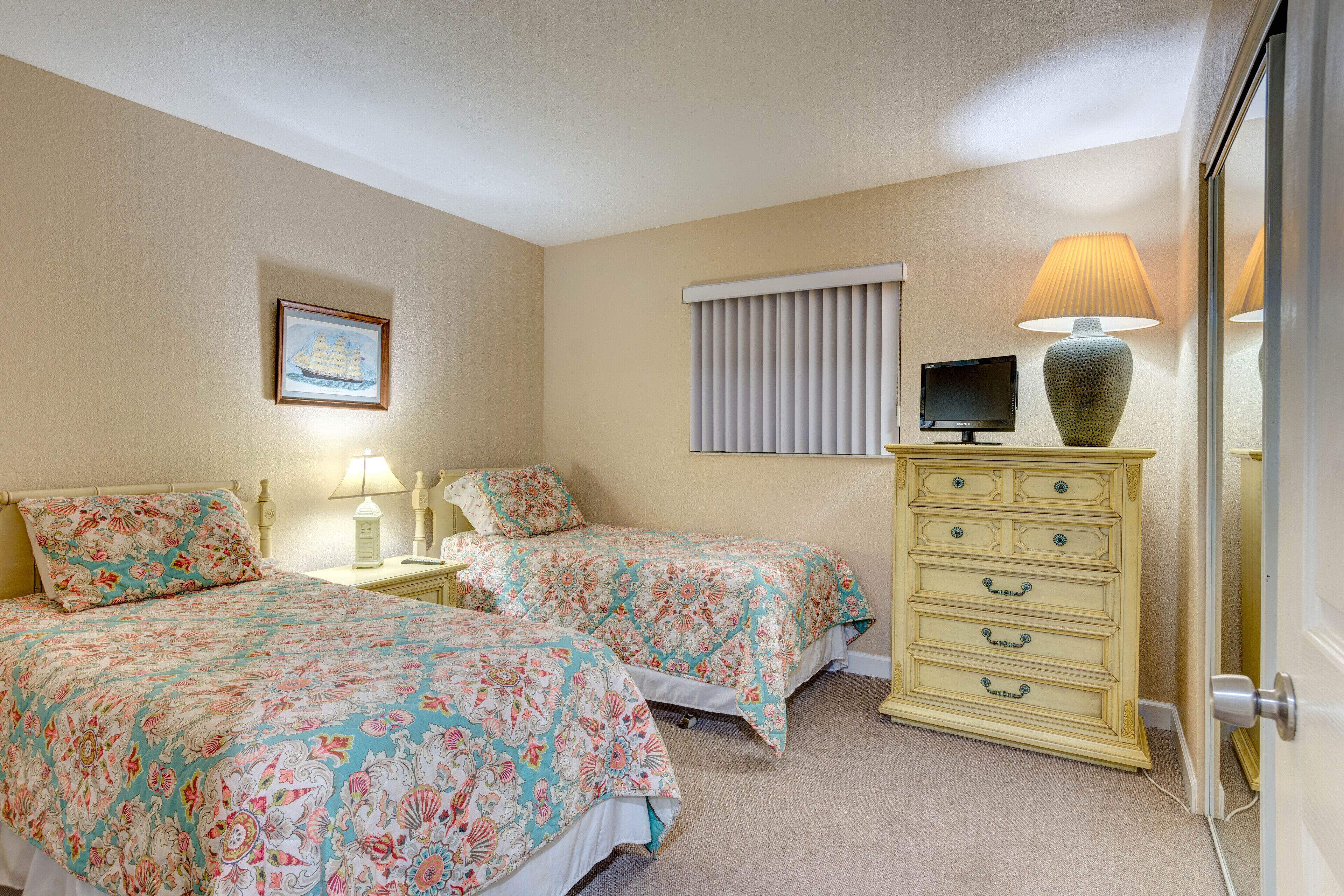 A bedroom with two beds, a dresser, a television, and a window with blinds.