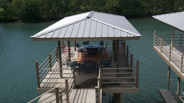 Un muelle con zona de estar cubierta y una mesa, rodeado de agua y árboles.