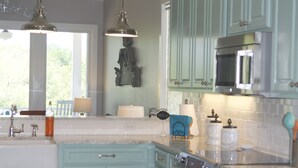 Una cocina con armarios azul verdoso, electrodomésticos de acero inoxidable, iluminación colgante y un amplio ventanal que proporciona luz natural.