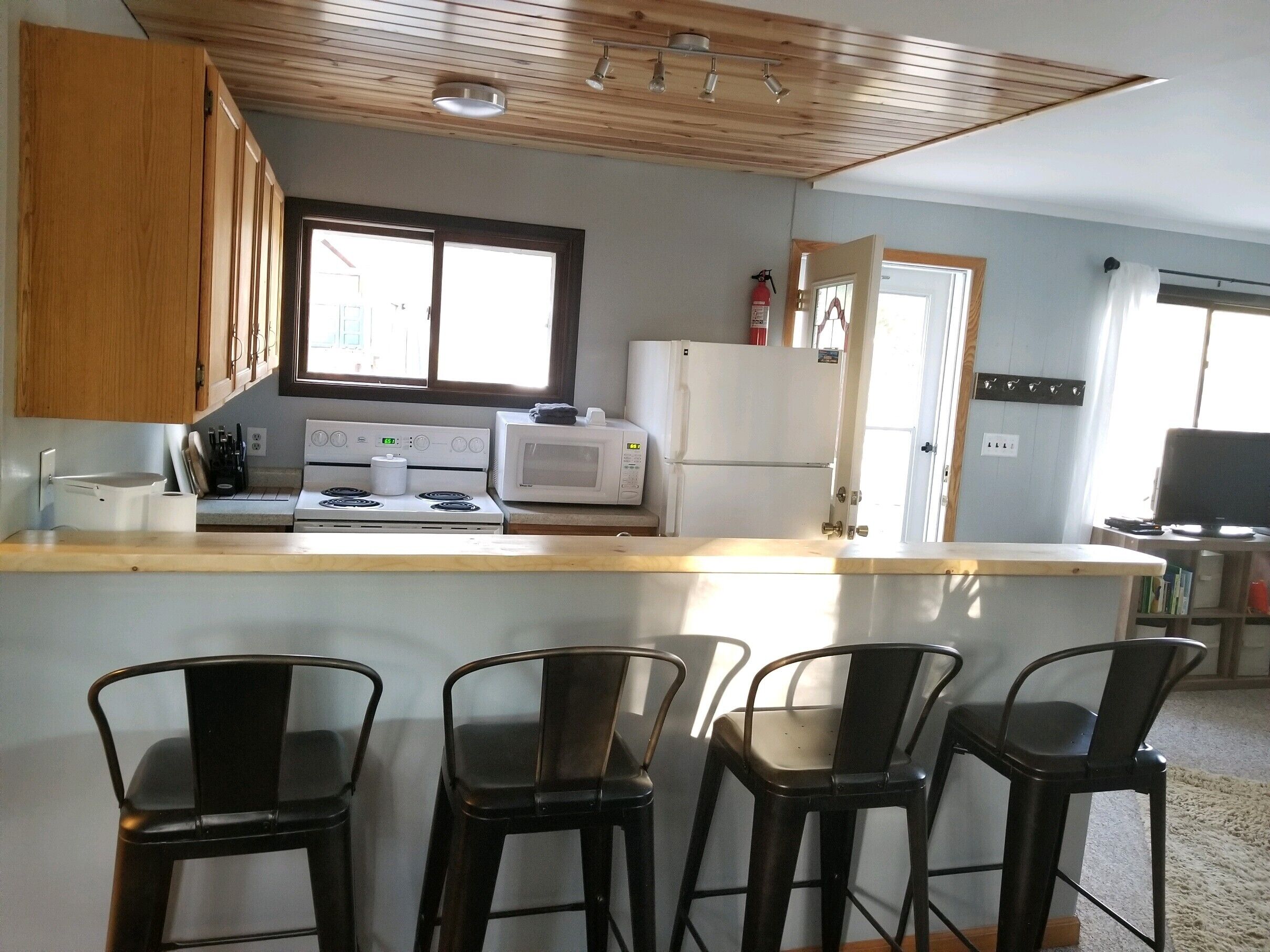 Una cocina con armarios de madera, estufa, microondas y refrigerador. Hay cuatro taburetes negros y un ventanal que proporciona luz natural.
