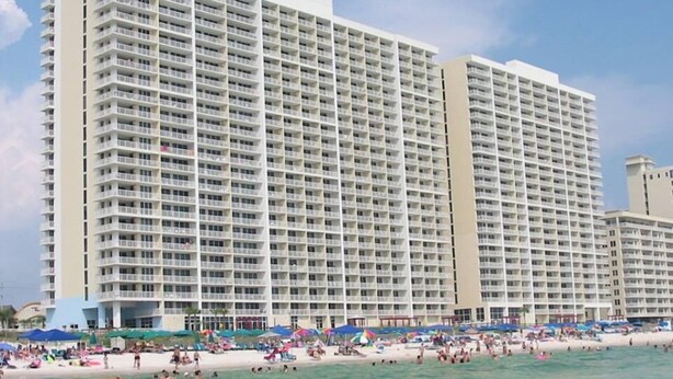 Een groot hotel met meerdere balkons, een strand op de voorgrond en een heldere lucht.