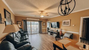 A living room with a black leather sofa, a wooden dining table, a ceiling fan, and a TV mounted on the wall. - 5 minutes to the beach! Cozy 2 King BR 2BA Condo at Myrtlewood Golf Club (Myrtle Beach)