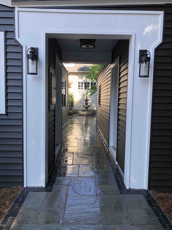 A narrow, wet stone pathway leading to a house entrance with a decorative fountain and two wall-mounted lanterns.