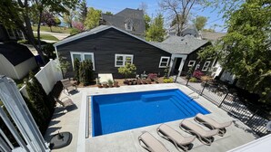A rectangular swimming pool with a clear blue water surface, surrounded by a concrete deck. There are four beige lounge chairs aligned along the pool's edge. The pool is adjacent to a house with a dark gray exterior and a sloped roof. - Lake Michigan Beach & Heated Pool 1 King  BR+  Cottage Sleeps 3 (South Haven)