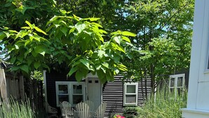 A garden pathway leading to a wooden deck with chairs, surrounded by lush greenery and flowering plants. - Lake Michigan Beach & Heated Pool 1 King  BR+  Cottage Sleeps 3 (South Haven)