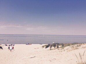 A beach with people walking and sitting on the sand, a clear sky, and the ocean in the background.
