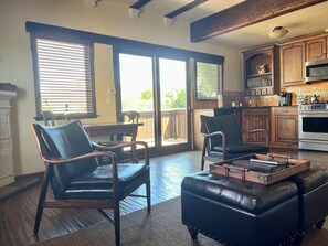 A spacious living area with a fireplace, a large window with blinds, a dining table, and a kitchen with wooden cabinets and a microwave.