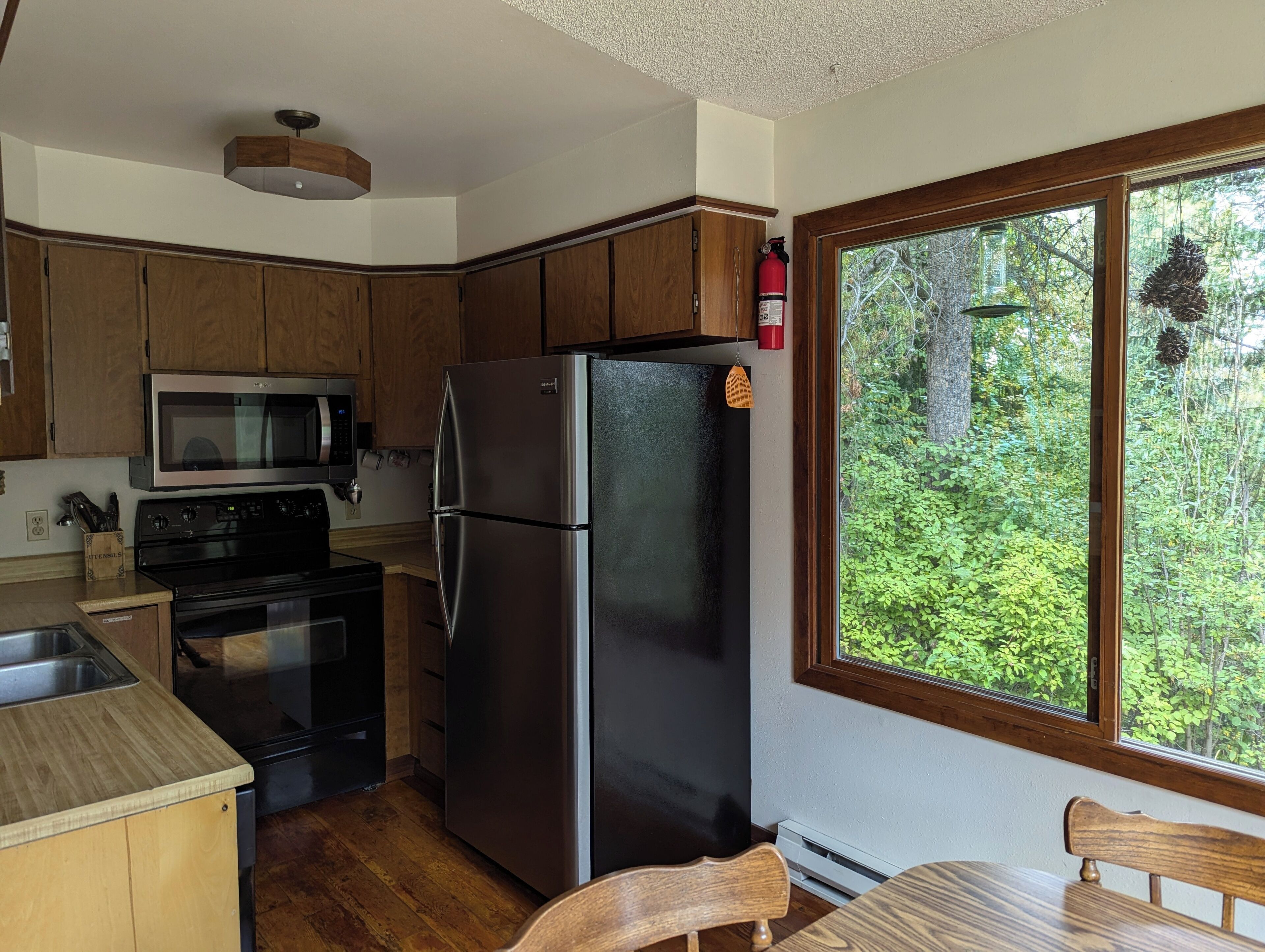 A compact kitchen with wooden cabinets, a stainless steel refrigerator, a microwave, and a stove. A window offers a view of greenery outside.