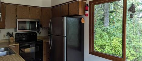 A compact kitchen with wooden cabinets, a stainless steel refrigerator, a microwave, and a stove. A window offers a view of greenery outside.
