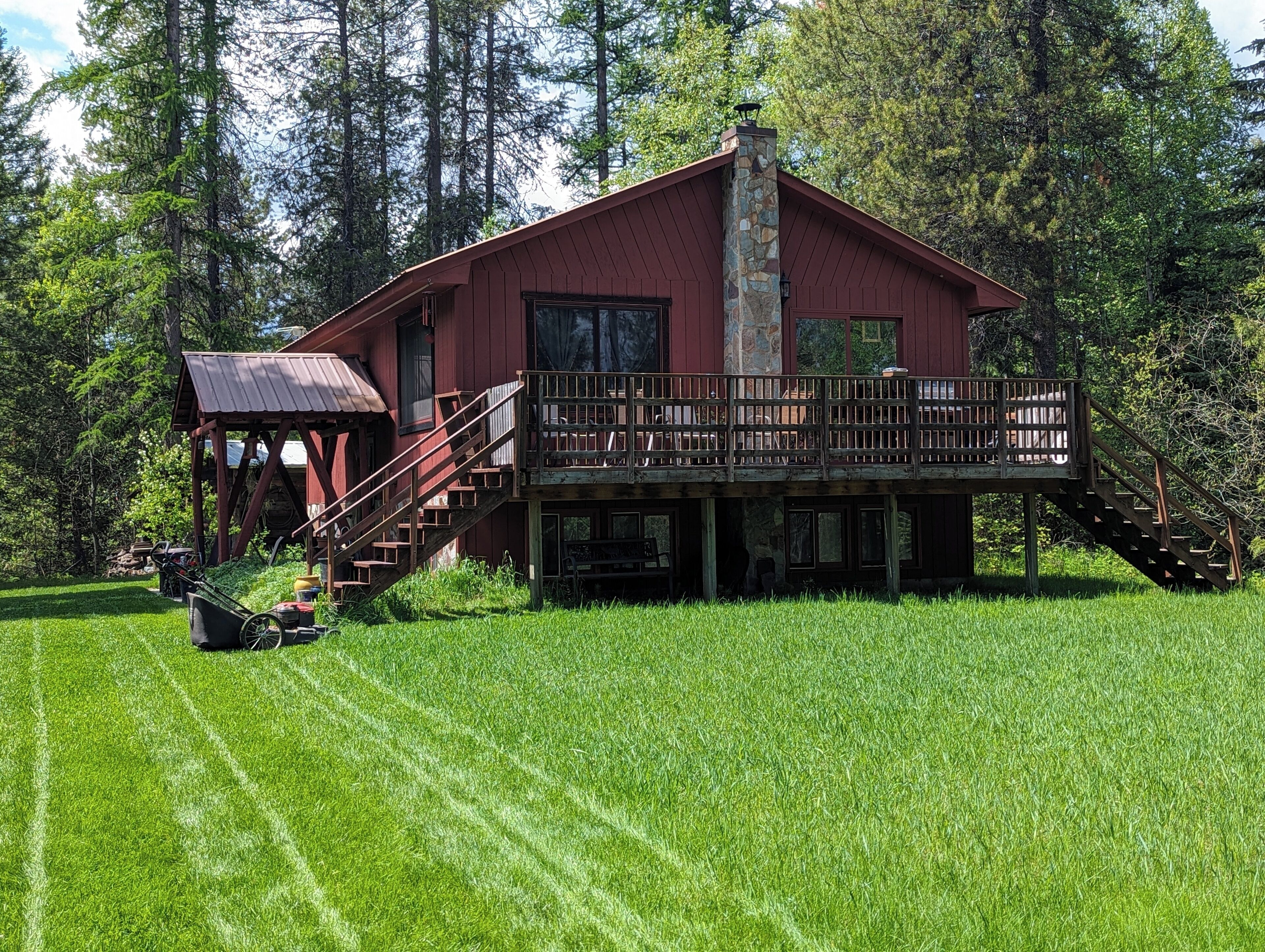 A red wooden cabin with a balcony and stairs, surrounded by a lush green lawn and tall trees.